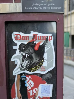 Street sticker The sticker showcases a black dog with a menacing expression, sharp teeth, and red horns. The dog wears a gold spiked collar. Above the dog, the text "Don Jump" is written in a gothic font. Below the dog, it is specified "Don Jump Tattoo". There is a second sticker under the first that contains a QR code and a profile name on Instagram.