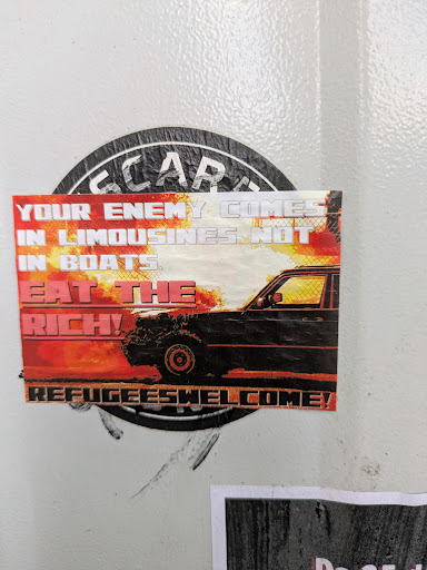 A rectangular sticker with a strong political message. It features the text 'YOUR ENEMY COMES IN LIMOUSINES NOT IN BOATS, EAT THE RICH! REFUGEES WELCOME!' over a background image of a black car against a fiery orange sky.