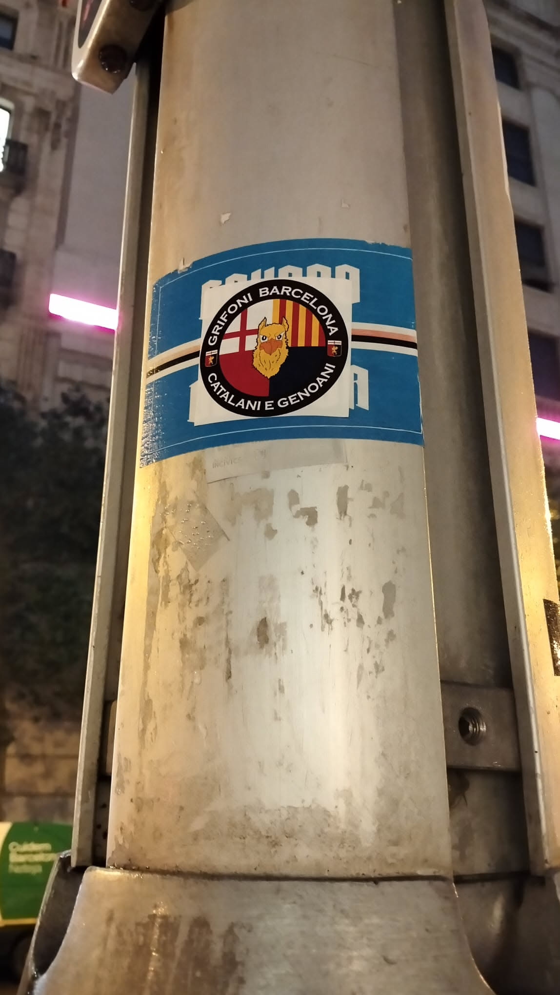 The sticker is predominantly blue and rectangular, featuring a central circular emblem. This emblem is divided vertically, with the left half displaying a red cross on a white background (St. George's Cross) and the right half showing the yellow and red vertical stripes of the Senyera flag. Superimposed on this divided background is a stylized, cartoonish griffin-like creature with a yellow body, orange face, and red beak. Around the circular emblem, white text reads 'GRIFONI BARCELONA' at the top and 'CATALANI E GENOANI' at the bottom. The blue rectangular background of the sticker has white and black horizontal stripes along its top and bottom edges, and a faint white outline of a castle or fortress is visible at the top. The sticker shows signs of wear and tear, including peeling edges and water marks.