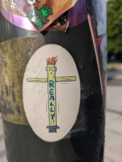 Street sticker An oval-shaped sticker featuring a simplistic, cartoon-style illustration of a cross-like figure. The vertical beam of the cross is depicted as a character with square-rimmed glasses and a wild tuft of orange hair or flames on its head. The word "REALLY!?" is prominently written in blue, capital letters vertically down the body of this character, ending with a question mark and exclamation mark. The background of the sticker is off-white, and it is adhered to a dark, textured pole.