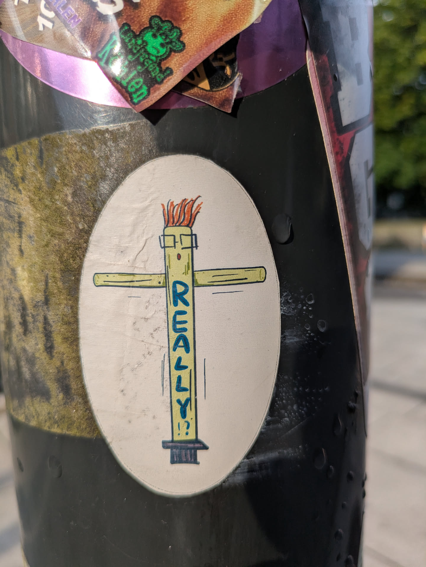 Street sticker An oval-shaped sticker featuring a simplistic, cartoon-style illustration of a cross-like figure. The vertical beam of the cross is depicted as a character with square-rimmed glasses and a wild tuft of orange hair or flames on its head. The word "REALLY!?" is prominently written in blue, capital letters vertically down the body of this character, ending with a question mark and exclamation mark. The background of the sticker is off-white, and it is adhered to a dark, textured pole.