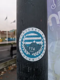 Street sticker The sticker is circular with a white border. The inner design features a stylized representation of mountains in dark blue, with white peaks. Above the mountains are horizontal blue and white stripes. Encircling the central design is a laurel wreath in white. Within the wreath, the number '116' is prominently displayed in white, with 'MMXIX' (Roman numerals for 2019) below it. The text around the outer edge of the sticker reads 'KVARTET GRABERGET BAJARNA' in white capital letters on a blue background.