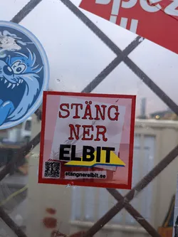 Street sticker The sticker is square-shaped with a red border. The background is a gradient of pink and white. The text 'STÄNG NER' is written in a stylized, bold red font. Below this, 'ELBIT' is written in large, bold black letters, with a yellow and blue graphic element underneath. A QR code is present on the left side of the sticker, and the website 'stängnerelbit.se' is printed below it. The sticker appears to be adhered to a reflective surface, possibly glass, with some scuff marks and reflections visible.