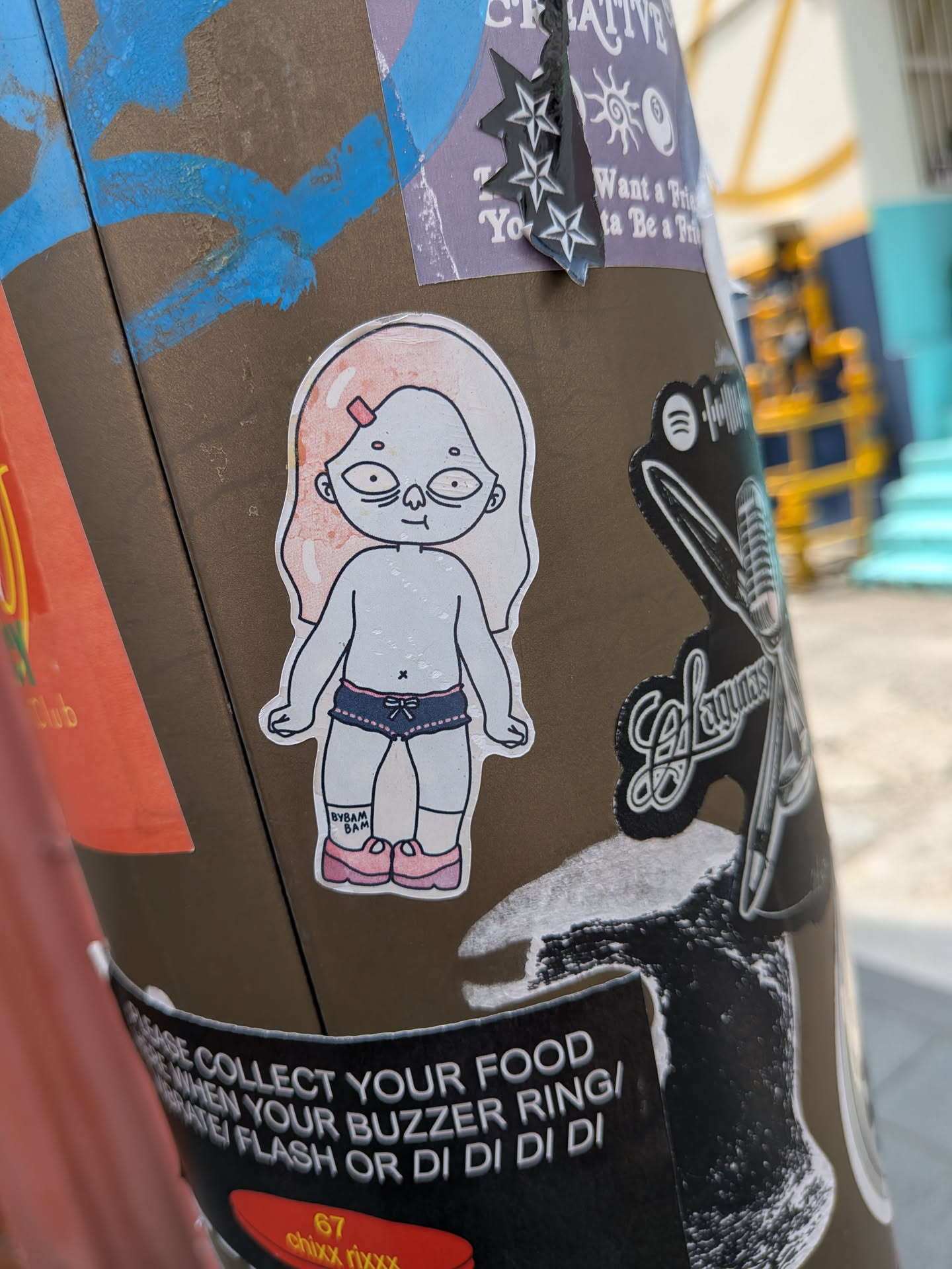 Die-cut sticker depicting a pale cartoon figure with large eyes, light pink hair, dark blue shorts, and pink shoes. The text 'BYBAM BAM' is written on the right ankle.