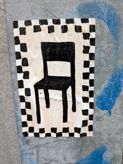 Street sticker The sticker features a black silhouette of a simple chair with a square seat and back, and four straight legs. The chair is rendered in a bold, thick black outline. It is set against a cream-colored background that appears to be paper. Surrounding the central image is a border of alternating black and white squares, resembling a checkerboard pattern. The paper has visible creases and wrinkles, indicating it is a physical sticker applied to a surface. The overall aesthetic is graphic and minimalist.