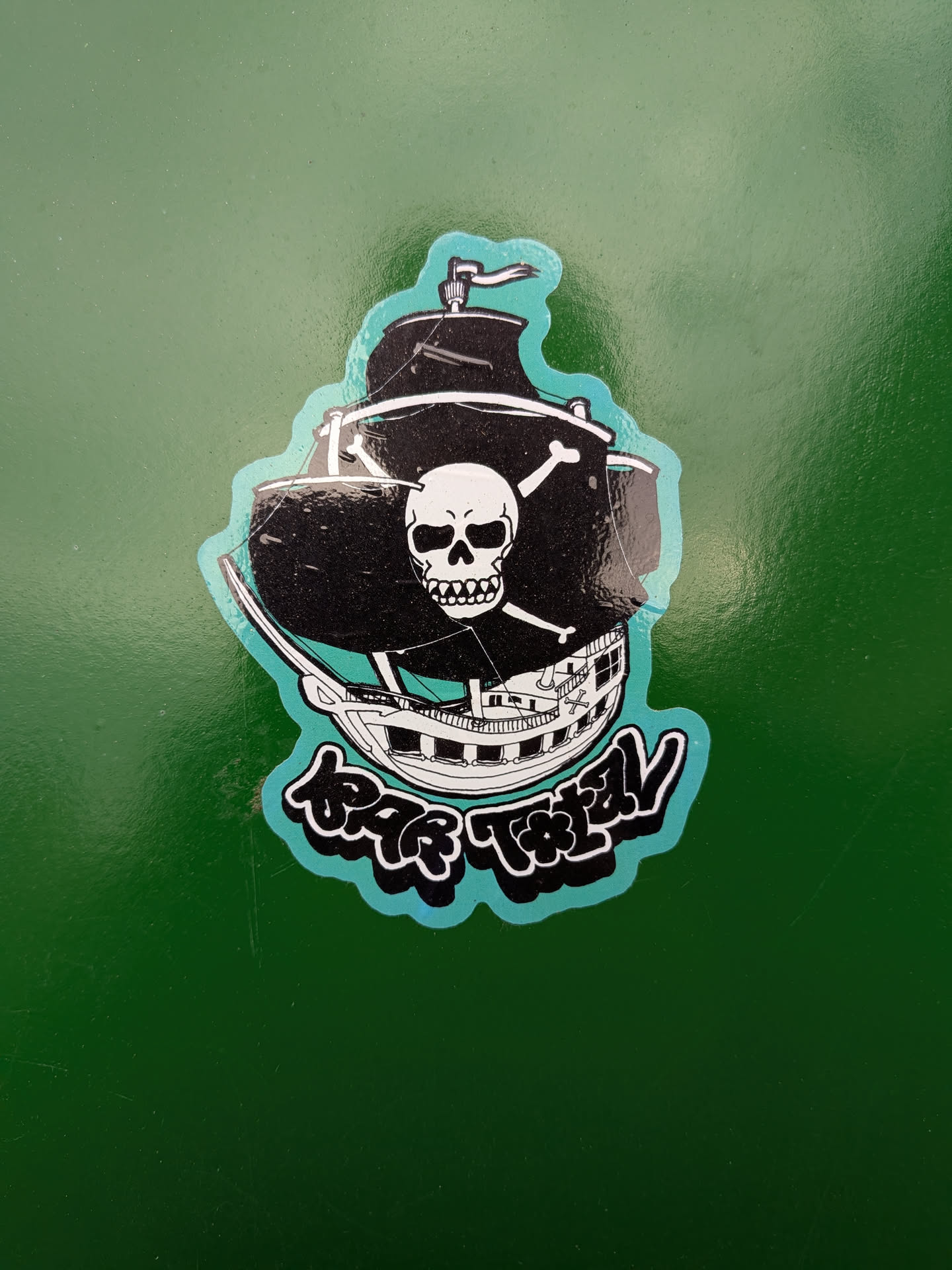 The sticker depicts a black pirate ship with white sails and rigging. A prominent skull and crossbones symbol is emblazoned on the main sail. The ship has multiple decks and windows, rendered in white and black outlines. Below the ship, there is stylized text in a bubble-like font with a turquoise outline and black fill, which appears to be 'BAR TOTAL'. The entire design is bordered by a wavy turquoise line.