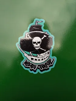 Street sticker The sticker depicts a black pirate ship with white sails and rigging. A prominent skull and crossbones symbol is emblazoned on the main sail. The ship has multiple decks and windows, rendered in white and black outlines. Below the ship, there is stylized text in a bubble-like font with a turquoise outline and black fill, which appears to be 'BAR TOTAL'. The entire design is bordered by a wavy turquoise line.