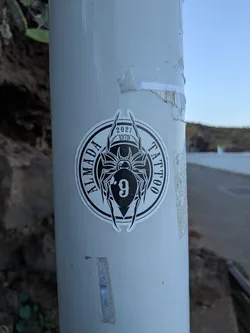 Street sticker A circular, die-cut sticker in black and white. It depicts a stylized spider with the number '9' on its abdomen. The text 'ALMADA TATTOO' arches around the sides, with '2021 ESTD' at the top.