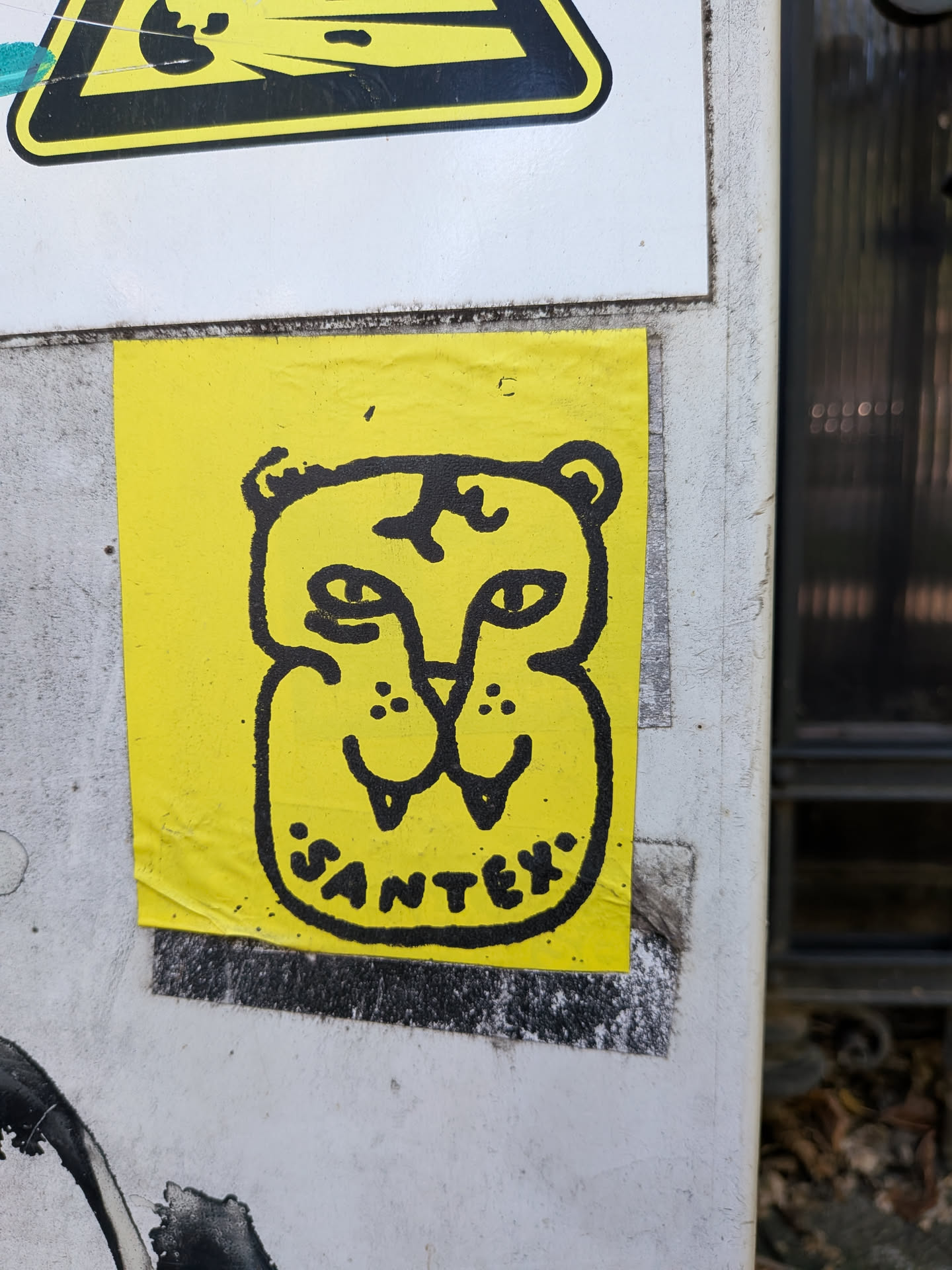 A rectangular yellow sticker featuring a thick black line drawing of a stylized tiger head. The character has fangs, small ears, and the word 'SANTEX' written along the bottom.