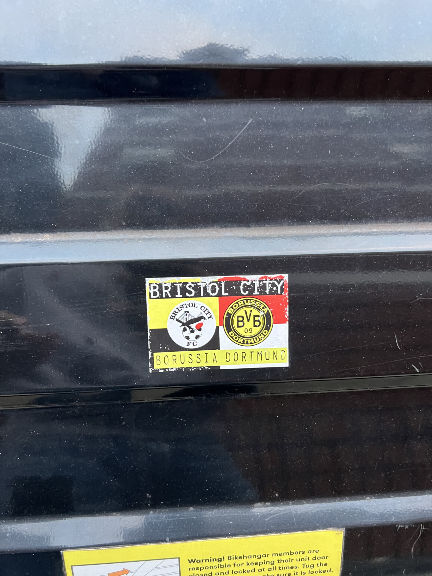 A rectangular sticker featuring a combined design of two football clubs. The sticker has a slightly worn appearance with minor scuffs and dust. The top left section displays 'BRISTOL CITY' in black text on a white background. Below this, the left half of the sticker features the Bristol City FC crest, which is a black circle with 'BRISTOL CITY' text, a robin bird, and 'FC' below it, all set against a background of yellow and black vertical stripes. The right half of the sticker showcases the Borussia Dortmund (BVB) logo, a yellow circle with 'BORUSSIA BVB 09 DORTMUND' text, placed on a background of red and black vertical stripes. At the very bottom of the sticker, 'BORUSSIA DORTMUND' is written in black text on a yellow background. The overall color scheme is dominated by yellow, black, white, and red.