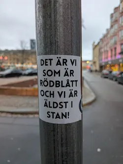Street sticker The sticker is rectangular with rounded corners and is predominantly white with black text. The text is arranged in several lines, with the largest font size on the first and third lines. The text reads 'DET ÄR VI SOM ÄR RÖDBLÅTT OCH VI ÄR ÄLDST I STAN!'. There is a partial circular emblem on the right side of the sticker, appearing to be dark blue with a white outline, but it is mostly cut off by the edge of the sticker.