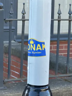 Street sticker The sticker is predominantly royal blue with bright yellow text and a yellow icon, outlined by a thin yellow border. Its shape is generally rectangular with a wavy, slightly curved top edge. The most prominent feature is the large, bold, yellow capital letters 'MONAK' in the center. Above the 'O' in 'MONAK', there is a yellow icon resembling a bell or a small bag/purse with a rounded top and a wider base. Below 'MONAK', in smaller yellow text, it reads 'Sticker Number', followed by the numerical identifier '02695'. The sticker appears to be a physical adhesive label, showing minor wear and slight peeling at the bottom edge, affixed to a light grey pole.