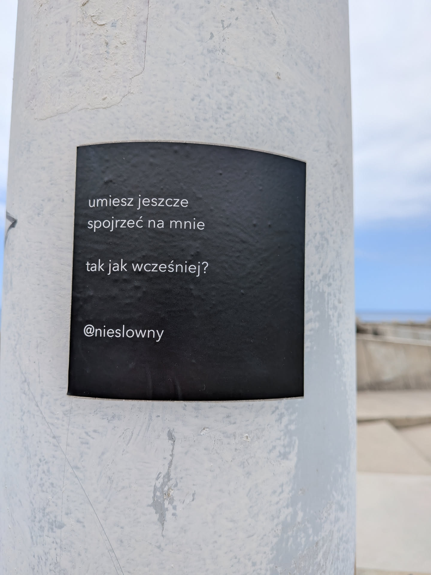 A square black sticker on a white pole featuring white sans-serif Polish text and a social media handle.