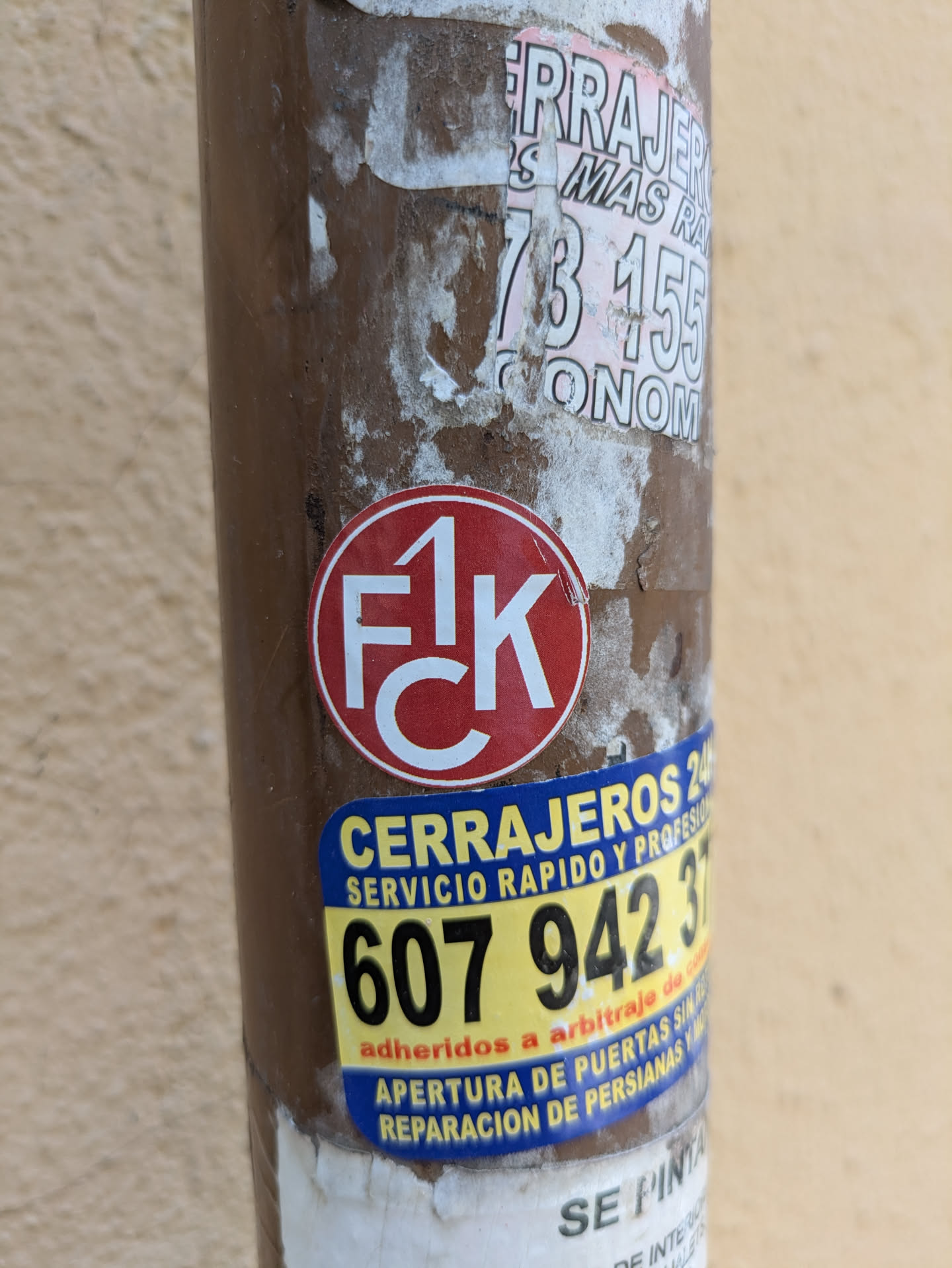 A weathered image shows a pole covered in various stickers. Centered is a red and white round sticker with "FCK" written inside. Below it, a yellow and blue sticker promotes locksmith services, displaying the phone number 607 942 37. The pole also has other partially torn stickers above the "FCK" sticker, with remnants of letters and numbers.