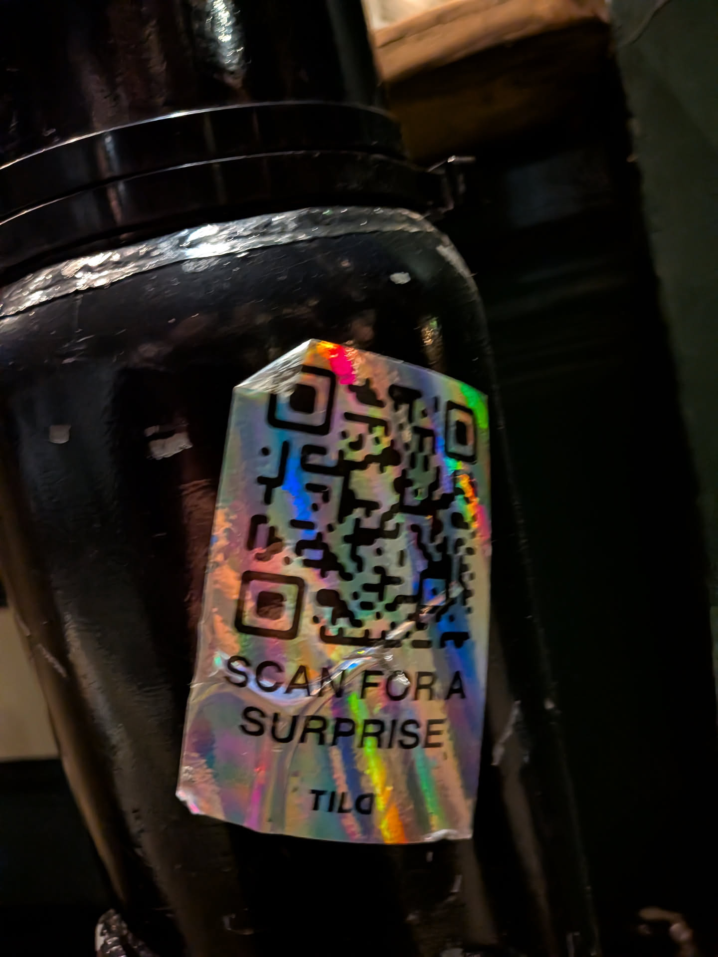 A rectangular, holographic sticker with an iridescent finish, reflecting rainbow colors. It features a prominent black QR code in the upper two-thirds. Below the QR code, the text 'SCAN FOR A SURPRISE' is printed in black, followed by 'TILO' at the very bottom. The sticker is applied to a dark, possibly black, cylindrical surface and shows signs of wear, including creases and slight peeling at the edges.