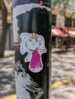 Street sticker A white sticker of a princess drawn with simple lines, wearing a crown and a pink dress, is adhered to a black metal pole. The drawing has a childish, playful style. There are other stickers above and below it. In the background, a blurry street scene unfolds.