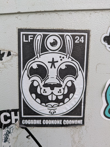 This black and white sticker showcases a cartoonish rabbit-like creature with long ears and a single eye located above its head. The creature has a wide grin, prominent teeth, and a star symbol on its forehead. The letters "LF" and the number "24" are printed in the upper corners, and the word "COONONE" is repeated along the bottom of the sticker.