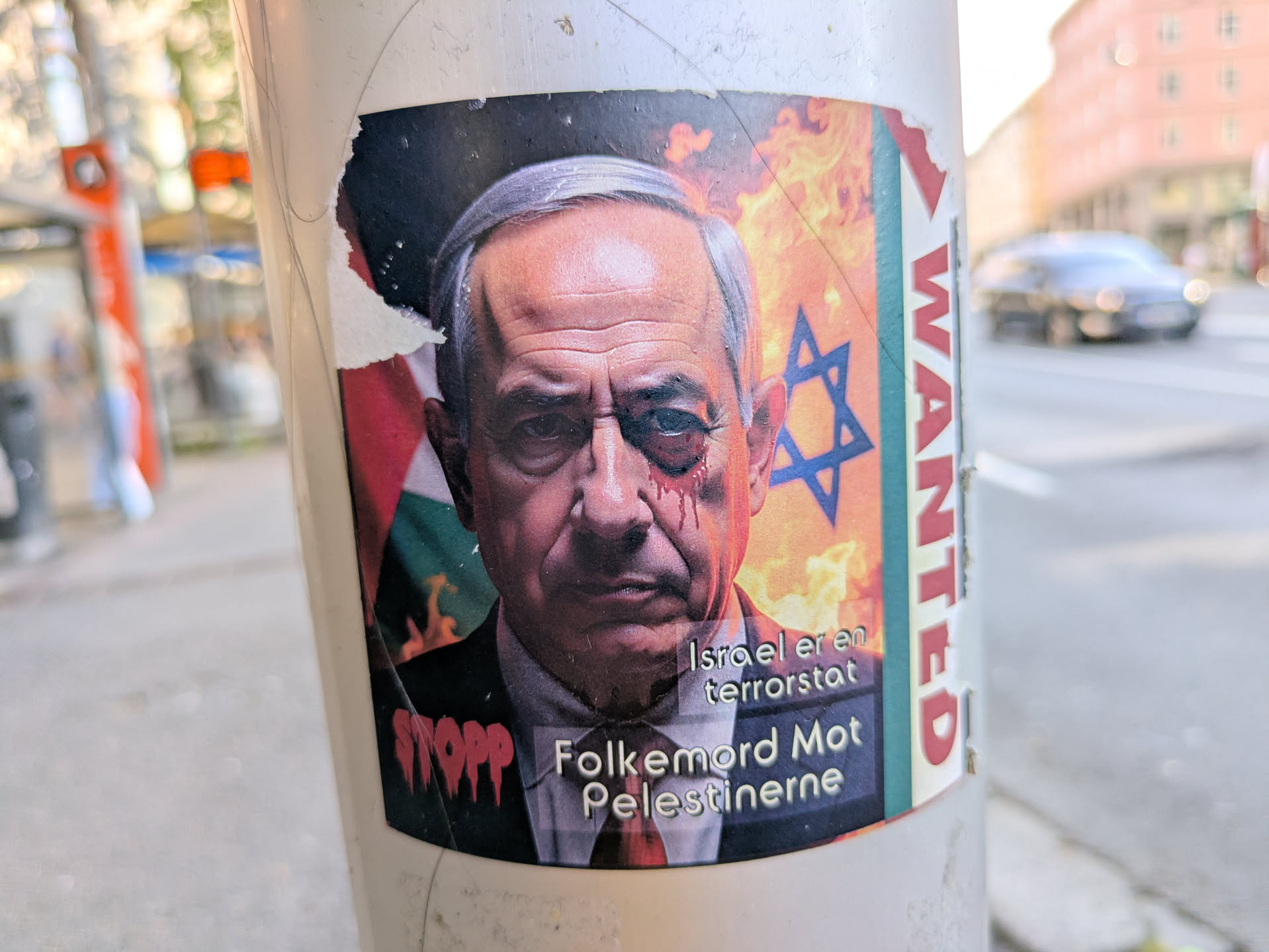 A politically charged sticker affixed to a white pole, depicting a solemn image of Benjamin Netanyahu with a bruised and bleeding left eye. The background on the left features a portion of the Palestinian flag, while the right side shows the Israeli Star of David flag superimposed over a fiery, orange background. Text on the sticker reads 'Israel er en terroristat' (Israel is a terrorist state) and 'Folkemord Mot Palestinerne' (Genocide Against Palestinians). The word 'STOPP' is visible vertically on the left edge and 'WANTED' on the right edge. The sticker expresses strong anti-Israel sentiment and solidarity with Palestinians.