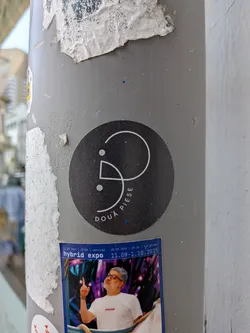 Street sticker A round black sticker affixed to a grey post. It displays a white abstract line logo with two dots and the curved Romanian text 'DOUĂ PIESE' along the bottom edge.