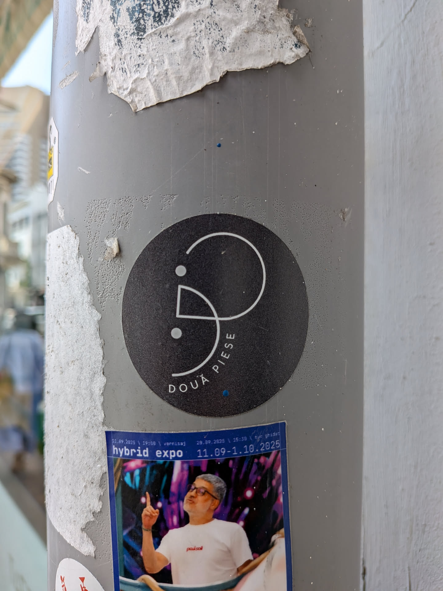 A round black sticker affixed to a grey post. It displays a white abstract line logo with two dots and the curved Romanian text 'DOUĂ PIESE' along the bottom edge.