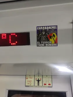 Street sticker A rectangular sticker with a dark purple background featuring three stylized figures. The central figure is a skull wearing a hooded jacket, with its hands clasped. Flanking the skull are two other hooded figures with masked faces. To the right of the central figure, there is a shield divided diagonally into yellow and red sections, bearing a black double-headed axe symbol. White block text at the top reads 'CARABANCHEL', followed by stylized white cursive text 'No Surrender'. At the bottom, white block text reads 'EL TERROR TIENE NOMBRE'. The overall aesthetic is gritty and rebellious.