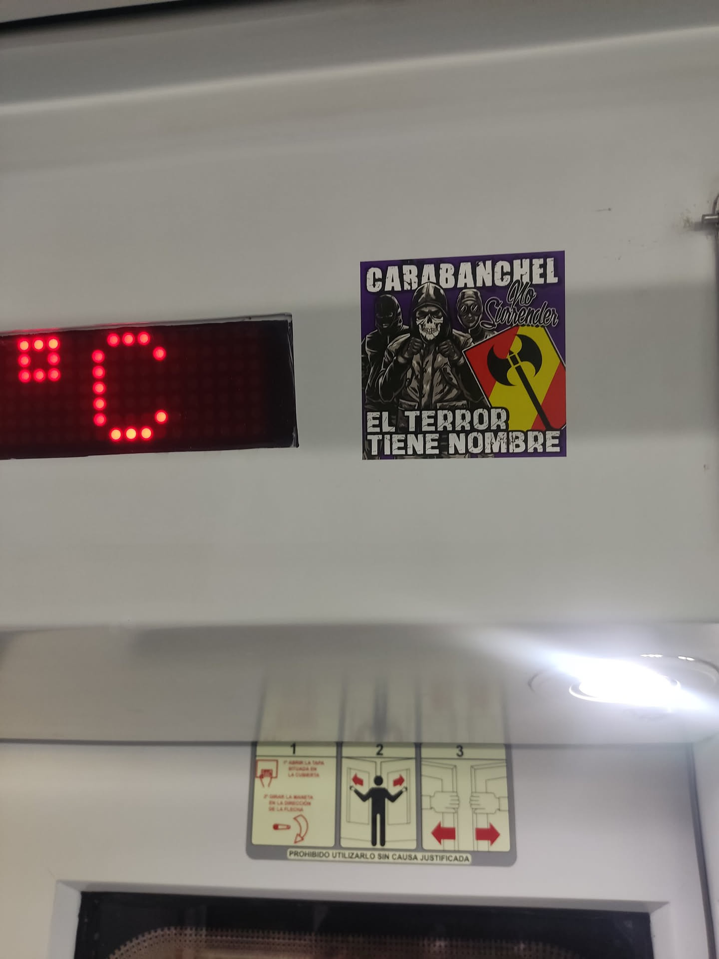 A rectangular sticker with a dark purple background featuring three stylized figures. The central figure is a skull wearing a hooded jacket, with its hands clasped. Flanking the skull are two other hooded figures with masked faces. To the right of the central figure, there is a shield divided diagonally into yellow and red sections, bearing a black double-headed axe symbol. White block text at the top reads 'CARABANCHEL', followed by stylized white cursive text 'No Surrender'. At the bottom, white block text reads 'EL TERROR TIENE NOMBRE'. The overall aesthetic is gritty and rebellious.