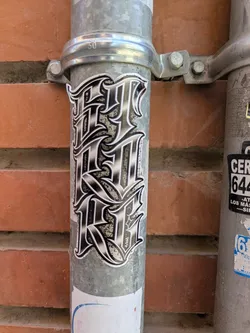 Street sticker The sticker features an elaborate, black and white design with the word "FOREVER" stacked vertically in a stylized font. It is affixed to a metallic pole, which is positioned against a brick wall. A small text near the design indicates "@naro.schiattuso".