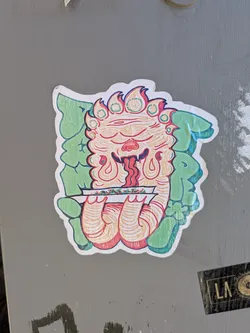 Street sticker The sticker features a stylized, cartoonish creature with a fiery mane and multiple eyes. Its face is predominantly orange and red, with a long, dripping red tongue. The creature is holding a rolled-up paper, possibly a joint, between its hands. Behind the creature are large, green, leafy shapes that form a backdrop. The overall style is illustrative and somewhat psychedelic, with bold outlines and vibrant colors.