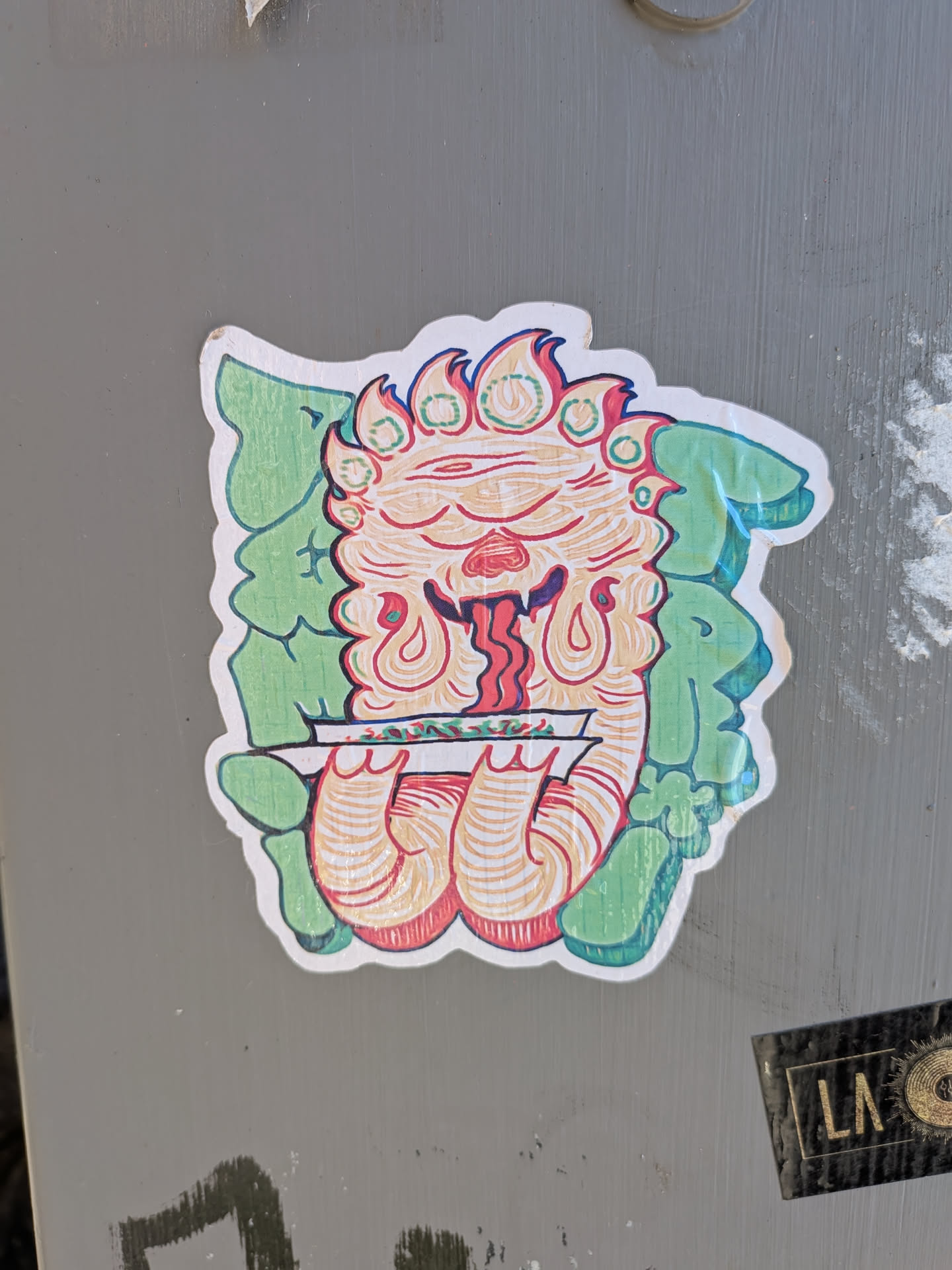 The sticker features a stylized, cartoonish creature with a fiery mane and multiple eyes. Its face is predominantly orange and red, with a long, dripping red tongue. The creature is holding a rolled-up paper, possibly a joint, between its hands. Behind the creature are large, green, leafy shapes that form a backdrop. The overall style is illustrative and somewhat psychedelic, with bold outlines and vibrant colors.