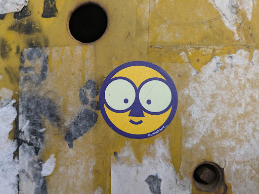 A round sticker features a yellow face with large, round, light green eyes and small black pupils. A dark blue nose and mouth complete the simple features. The face is outlined in dark blue. The sticker appears on a yellow, textured surface with some peeling paint.