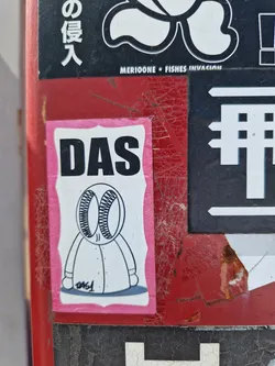 Street sticker A pink sticker featuring a simple illustration of a character with a das written above it. The character has a simplistic design and appears to be wearing a robe or coat. The number 0461 is visible near the character's feet. The sticker is affixed to a surface with other stickers and markings.