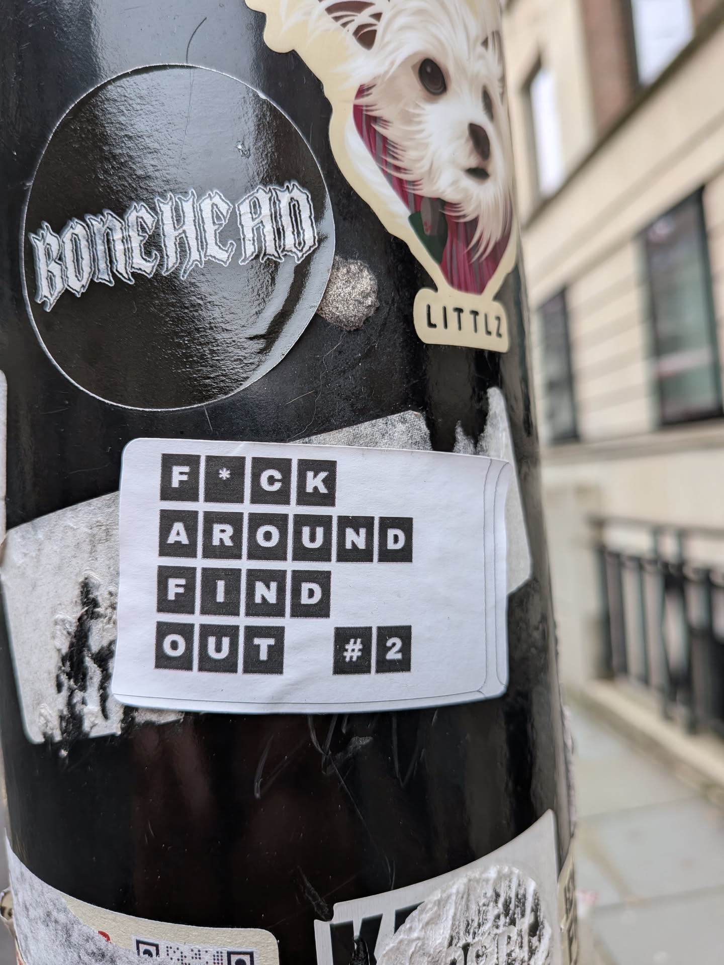 The sticker is rectangular with slightly rounded corners, featuring a white background. It displays text in a grid-like format, where each letter is a white character set within a black square. The text is arranged over four lines, reading 'F*CK', 'AROUND', 'FIND', and 'OUT #2'. The overall design is minimalist and bold, with a clear contrast between the black squares and white background/letters. The sticker appears slightly worn, with a small curl at the top right corner.