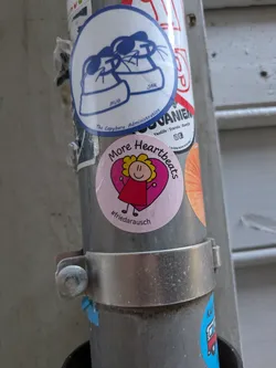 Street sticker The sticker is round and pink, displaying a cheerful cartoon character with blonde, curly hair and a red dress. This character is positioned in front of a prominent dark pink heart. Around the top edge of the circle, the words 'More Heartbeats' are written, and at the bottom, the hashtag '#friedarausch' is visible.