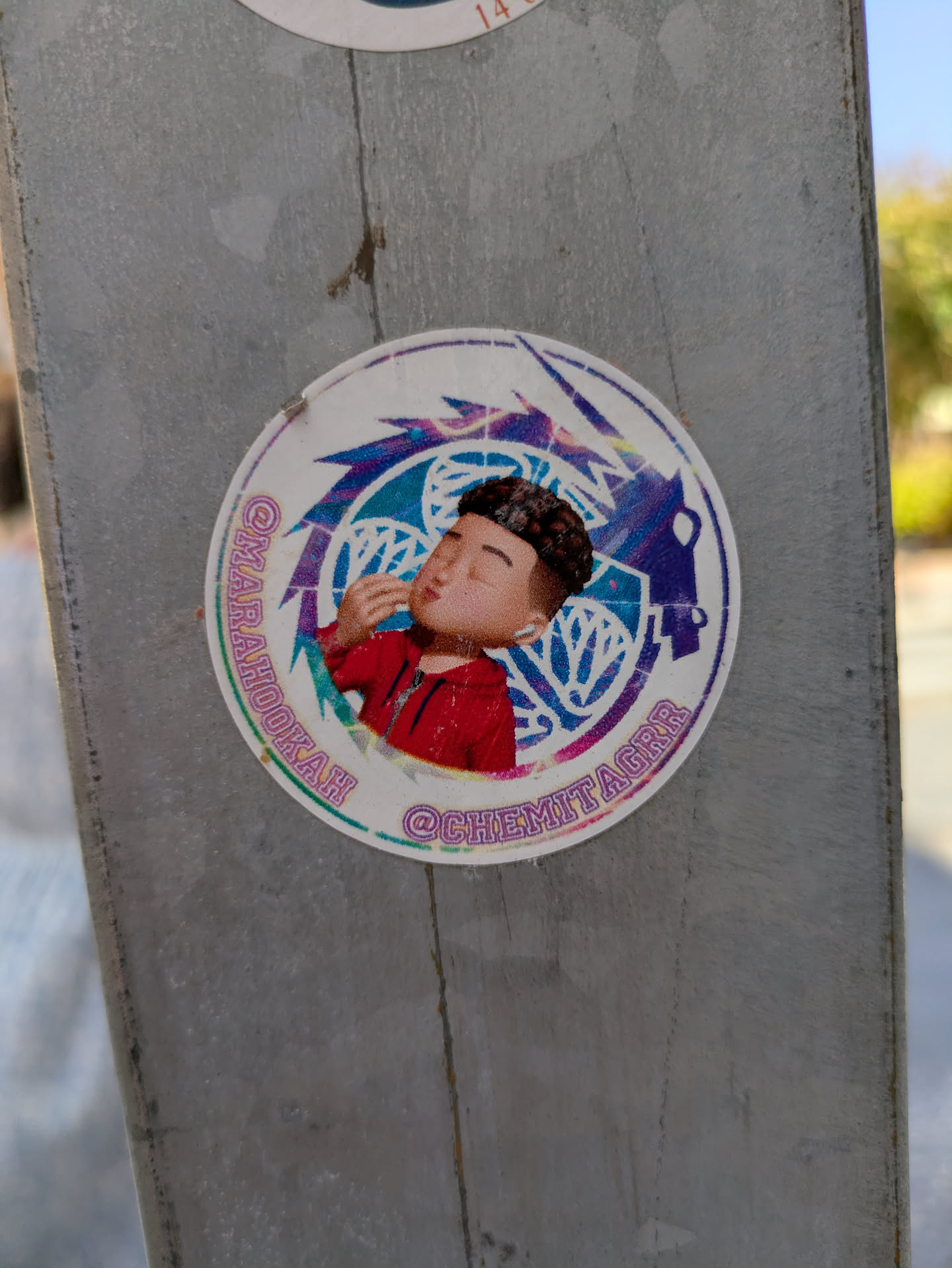 Street sticker This is a round sticker affixed to a gray metal surface. At the center, there is a cartoon-style drawing of a man with dark curly hair wearing a red jacket. He appears to be 'air kissing'. The background is a design with blue and purple colors, resembling wings or abstract shapes. Two usernames, @MARAHOOKAH and @CHEMITAGRR, are printed around the edge of the sticker. The overall style is vibrant, with a mix of colors and a slightly worn look.
