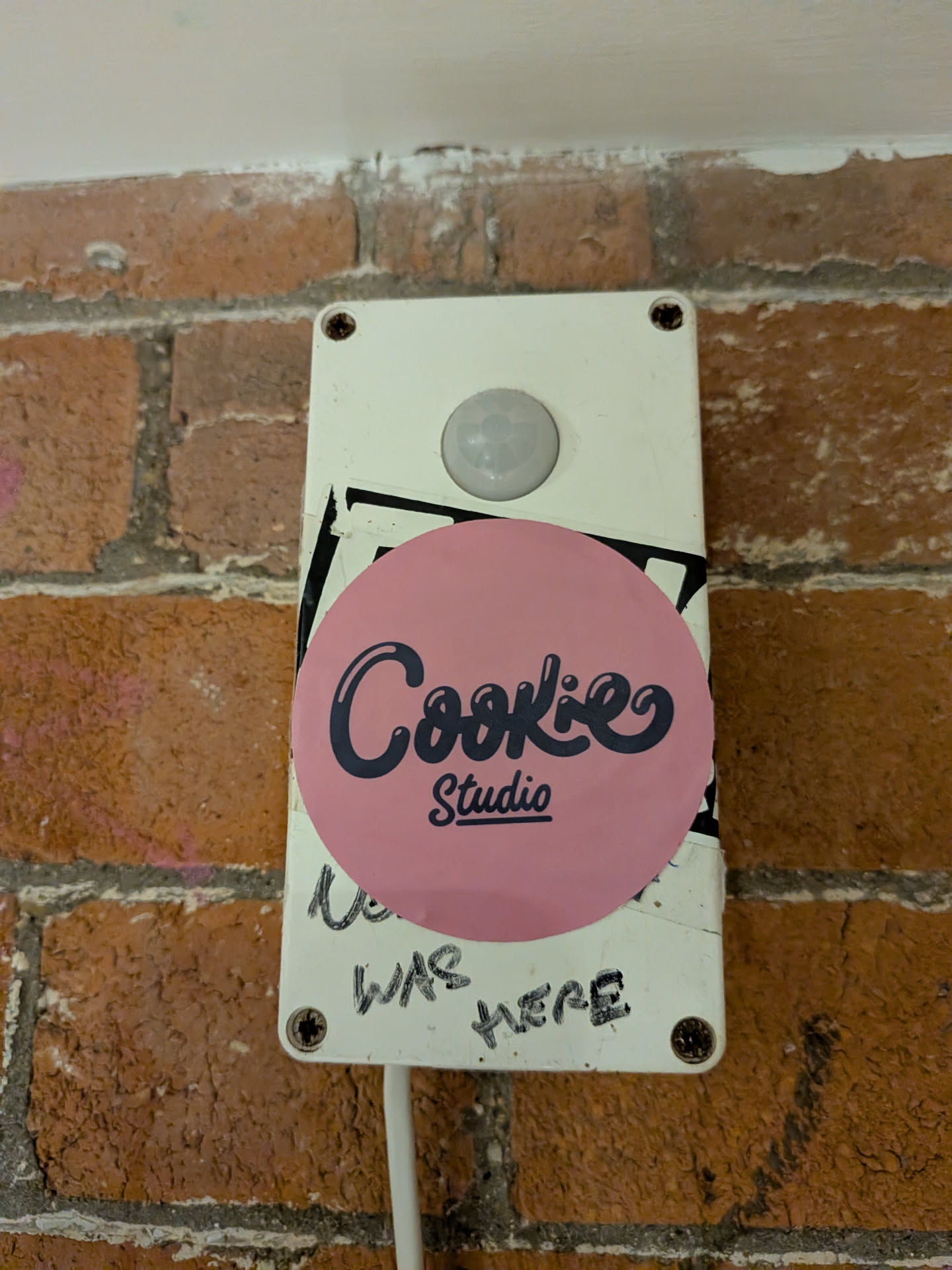 The sticker is circular with a vibrant, solid pink background. Centered on the pink circle is the text "Cookie" in a stylized, dark blue or black script font, featuring a subtle bubble or outline effect. Below "Cookie", in a smaller, sans-serif font, is the word "Studio", also in dark blue or black. The sticker appears glossy, showing some reflections, and is affixed to a white rectangular utility box, partially overlapping a black sticker underneath.