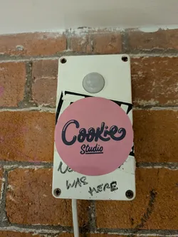 Street sticker The sticker is circular with a vibrant, solid pink background. Centered on the pink circle is the text "Cookie" in a stylized, dark blue or black script font, featuring a subtle bubble or outline effect. Below "Cookie", in a smaller, sans-serif font, is the word "Studio", also in dark blue or black. The sticker appears glossy, showing some reflections, and is affixed to a white rectangular utility box, partially overlapping a black sticker underneath.