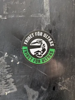 Street sticker The sticker is circular with a black border. The top half of the border contains white text that reads 'FRIHET FÖR ULTRAS' in a bold, sans-serif font. Inside this text, there is a white circle containing a black silhouette of a clenched fist holding a baton or club. The bottom half of the border is a vibrant green, also containing white text that reads 'FRIHET FÖR ULTRAS' in the same font. The overall design is stark and impactful, using a limited color palette of black, white, and green.