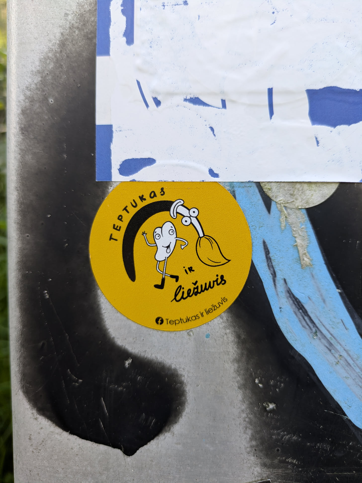 A vibrant yellow circular sticker is affixed to a metal pole, which is partly covered in black spray paint. The sticker prominently displays two whimsical cartoon characters: one is a white, blob-like figure with large eyes, waving, and wearing boots, possibly representing a tongue or a paint splash. The other character is a white paintbrush with eyes, holding a paint palette. The phrase 'TEPTUKAS IR LIEŽUVIS' is written in black text, curving along the top edge of the sticker. Below the characters, the same phrase 'Teptukas ir liežuvis' appears in smaller font, accompanied by a Facebook icon, indicating a social media presence.