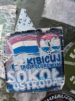 Street sticker The sticker is worn and creased, displaying a vibrant design despite its age. The top portion features a horizontal tricolor flag (red, white, blue) alongside a circular emblem. This emblem showcases a white eagle with outstretched wings clutching a soccer ball, set against a red, white, and blue background with white stars. Below this, the text 'KIBICUJ TRÓJKOLOROWYM SOKÓŁ OSTRÓDA' is prominently displayed in blue and white. Two small cartoon figures, a boy and a girl holding flags, are subtly visible in the background of the lower text section. The overall aesthetic is one of sports fandom and regional pride.