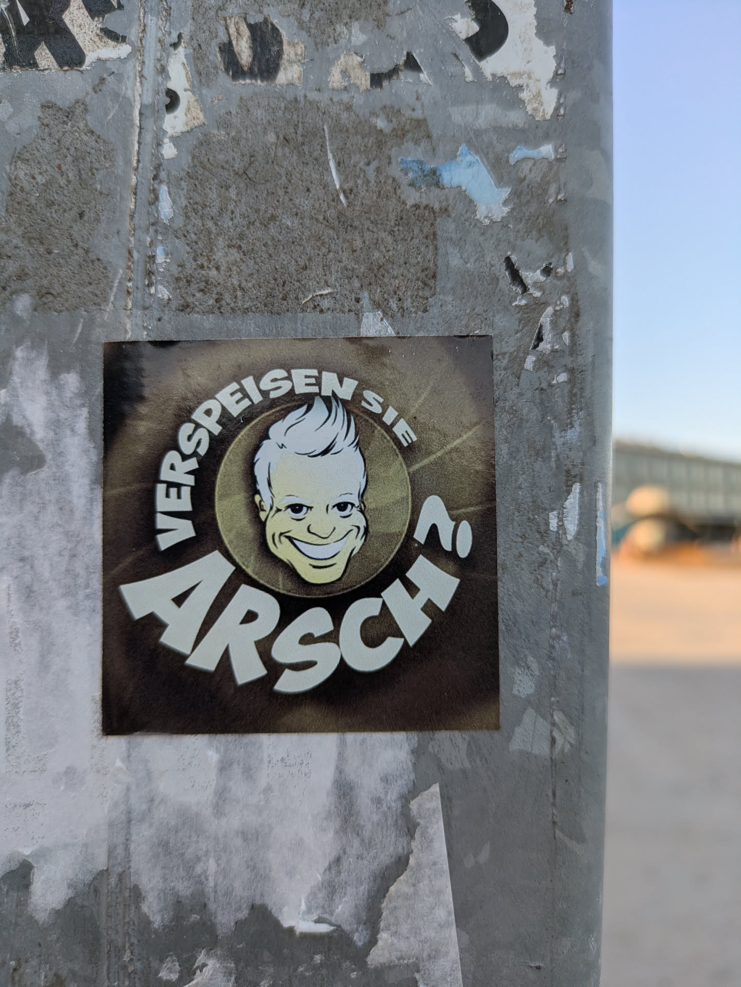 The sticker features a cartoonish face with a sly grin, surrounded by the text "Verspeisen Sie Arsch?". The sticker is adhered to a metal pole, which is covered in remnants of other stickers. The phrase translates to "Eat Ass?".