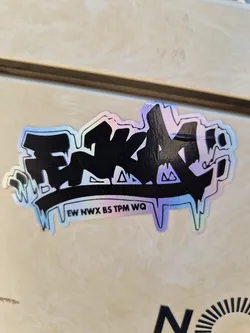 A sticker with a black graffiti-style design on an iridescent background. The text 'EW NWX BS TPM WQ' is visible at the bottom. 