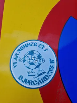 Street sticker A round sticker featuring a cartoon soccer ball character with the text "Vi är Nummer Ett Djurgårdens IF" which translates to "We are number one Djurgårdens IF".  The sticker is affixed to a surface with yellow, red, and blue sections.