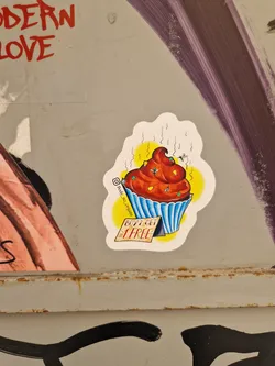 A sticker featuring an illustration of a cupcake made of poop, with flies around it. It has a yellow background and a small tag that says "Buy 2 Get 1 Free".  The sticker has a stylized design, and the overall aesthetic is humorous and slightly edgy.