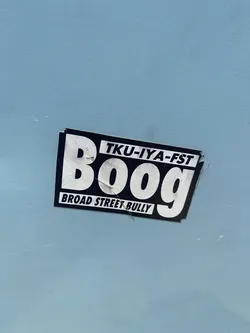 Street sticker The sticker is rectangular with a black border and a white background. The word 'Boog' is prominently displayed in large, bold, white sans-serif letters. Above 'Boog', in smaller white text, is 'TKU-IYA-FST'. Below 'Boog', also in smaller white text, is 'BROAD STREET BULLY'. The sticker appears slightly worn and has a few small tears at the edges.