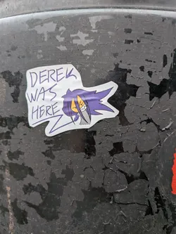 Street sticker The sticker features a stylized cartoon character with vibrant purple, spiky hair that extends sharply to the right. The character has yellow, almond-shaped eyes with black pupils and an angry, determined expression, showing sharp white teeth. A grey, almost metallic-looking element is visible below the character's face, possibly part of a collar or clothing. To the left of the character, in a handwritten, somewhat jagged purple font, are the words 'DEREK WAS HERE'. The entire design is outlined with a clean white border, giving it a distinct cut-out appearance against the dark, peeling background surface.