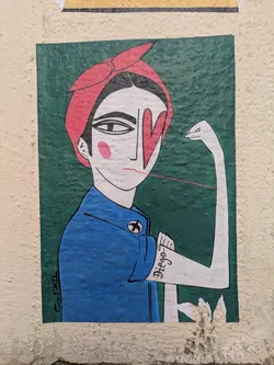 Street sticker The sticker shows a woman with a red headscarf and a blue shirt, reminiscent of Rosie the Riveter. However, the artistic style is unique. Instead of a nose, she has a red heart shape covering her right eye. She is flexing her arm and has a tattoo that reads "Diego" on her bicep. The background is a textured green, and the style is reminiscent of painted poster art.