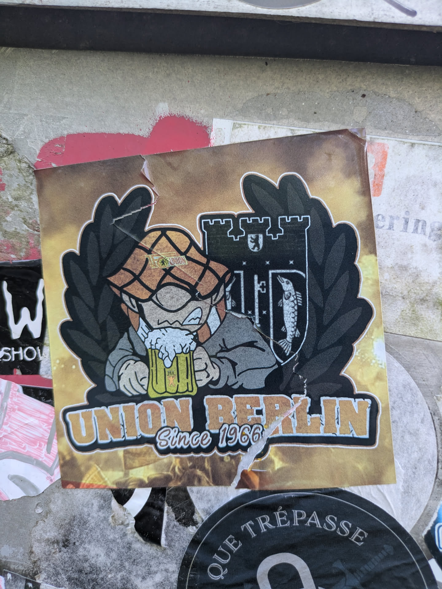 The sticker displays a cartoonish character with a checkered hat and a grey jacket, eagerly drinking from a frothy yellow beer mug. Behind the character is a dark, shield-like emblem framed by a dark wreath. The emblem features a castle silhouette on the left, containing a small bear, and a stylized fish on the right. Below the central image, the words 'UNION BERLIN' are prominently displayed in a gold-outlined font, with 'Since 1966' written underneath.