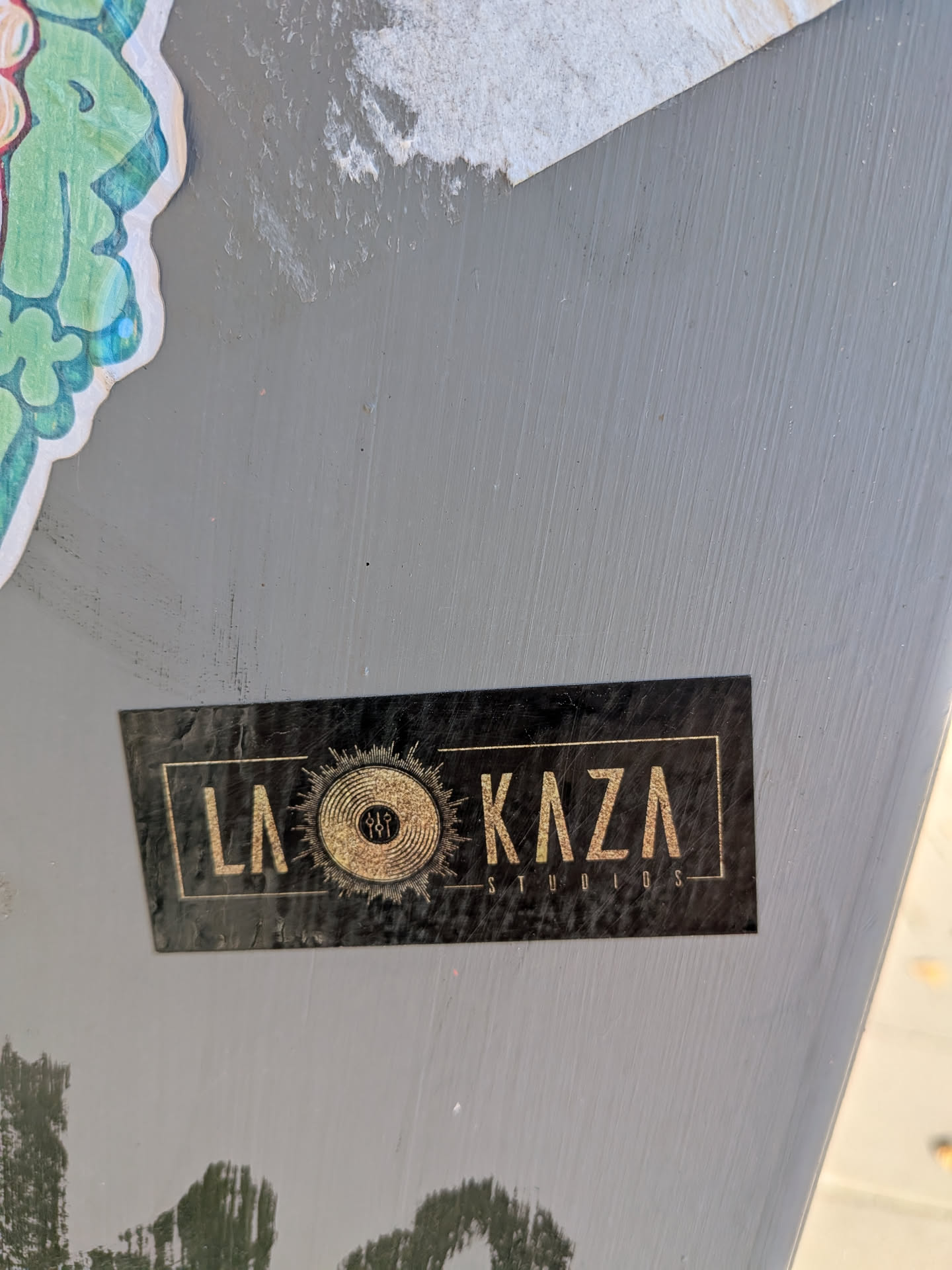 The sticker is rectangular with a black background and gold lettering. The text 'LA KAZA' is prominently displayed in a stylized font. To the right of 'LA', there is a circular emblem resembling a vinyl record with radiating lines, suggesting a sun or a sound wave. Inside the vinyl record graphic, there are three small figures. Below 'KAZA', the word 'STUDIOS' is written in a smaller, sans-serif font. The sticker has a slightly worn or textured appearance, indicating it has been applied.