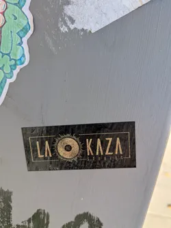 Street sticker The sticker is rectangular with a black background and gold lettering. The text 'LA KAZA' is prominently displayed in a stylized font. To the right of 'LA', there is a circular emblem resembling a vinyl record with radiating lines, suggesting a sun or a sound wave. Inside the vinyl record graphic, there are three small figures. Below 'KAZA', the word 'STUDIOS' is written in a smaller, sans-serif font. The sticker has a slightly worn or textured appearance, indicating it has been applied.