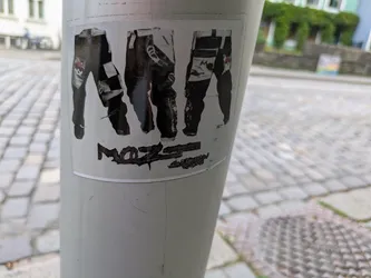 Street sticker The sticker displays three separate images of what appear to be motocross or racing pants, each with distinct designs. The leftmost pair shows a black and white checkered pattern and a red graphic, possibly a skull or logo, near the knee. The middle pair is predominantly dark with some lighter accents, and the rightmost pair also features a mix of dark and light sections with a small red detail. Beneath the pants, there is dark, distressed text; the first part is decipherable as 'MoZ', while the subsequent text is heavily worn and largely illegible. The sticker is applied to a light-colored utility pole set against a background of cobblestone pavement and distant buildings.